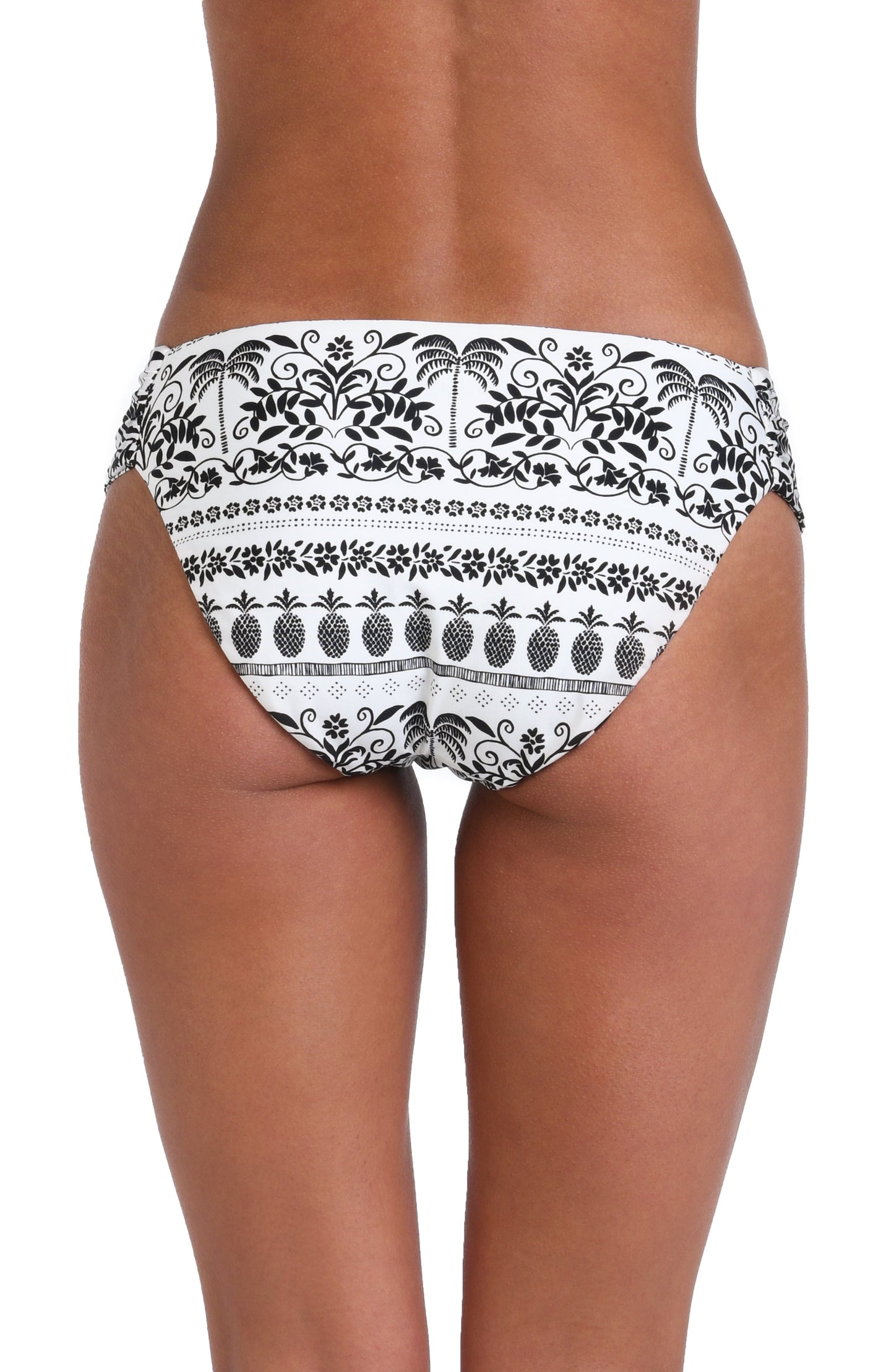 Model is wearing a white and black monochromatic patterned side shirred hipster bottom that features an allover tropical motif with hints of palm trees and pineapples set against a black background.